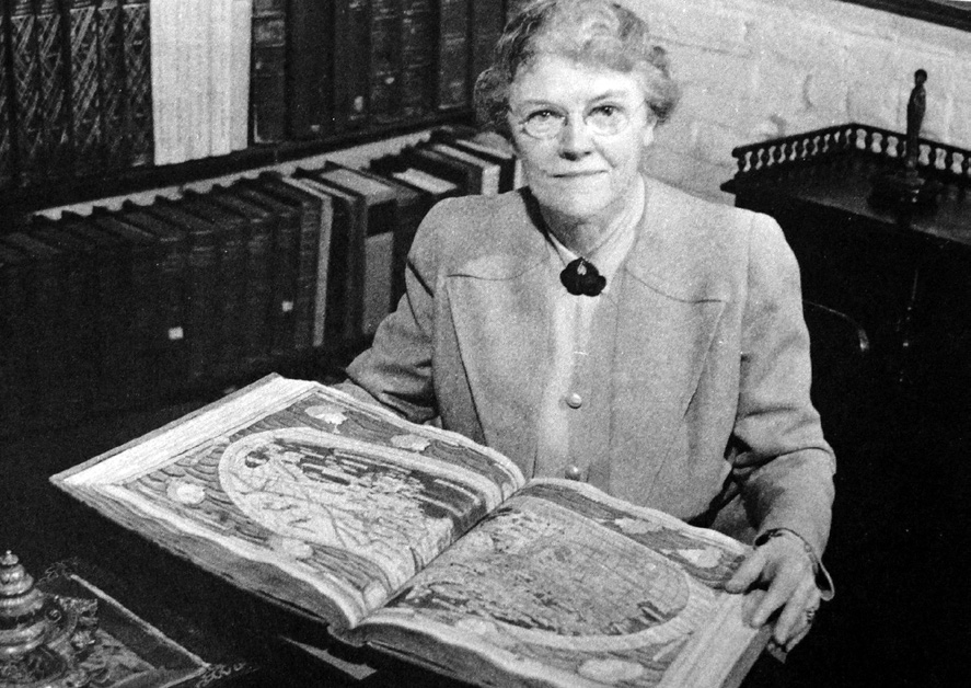 Black and white image of Margaret Stillwell sitting at a desk and holding an open illuminated manuscript in a library setting.