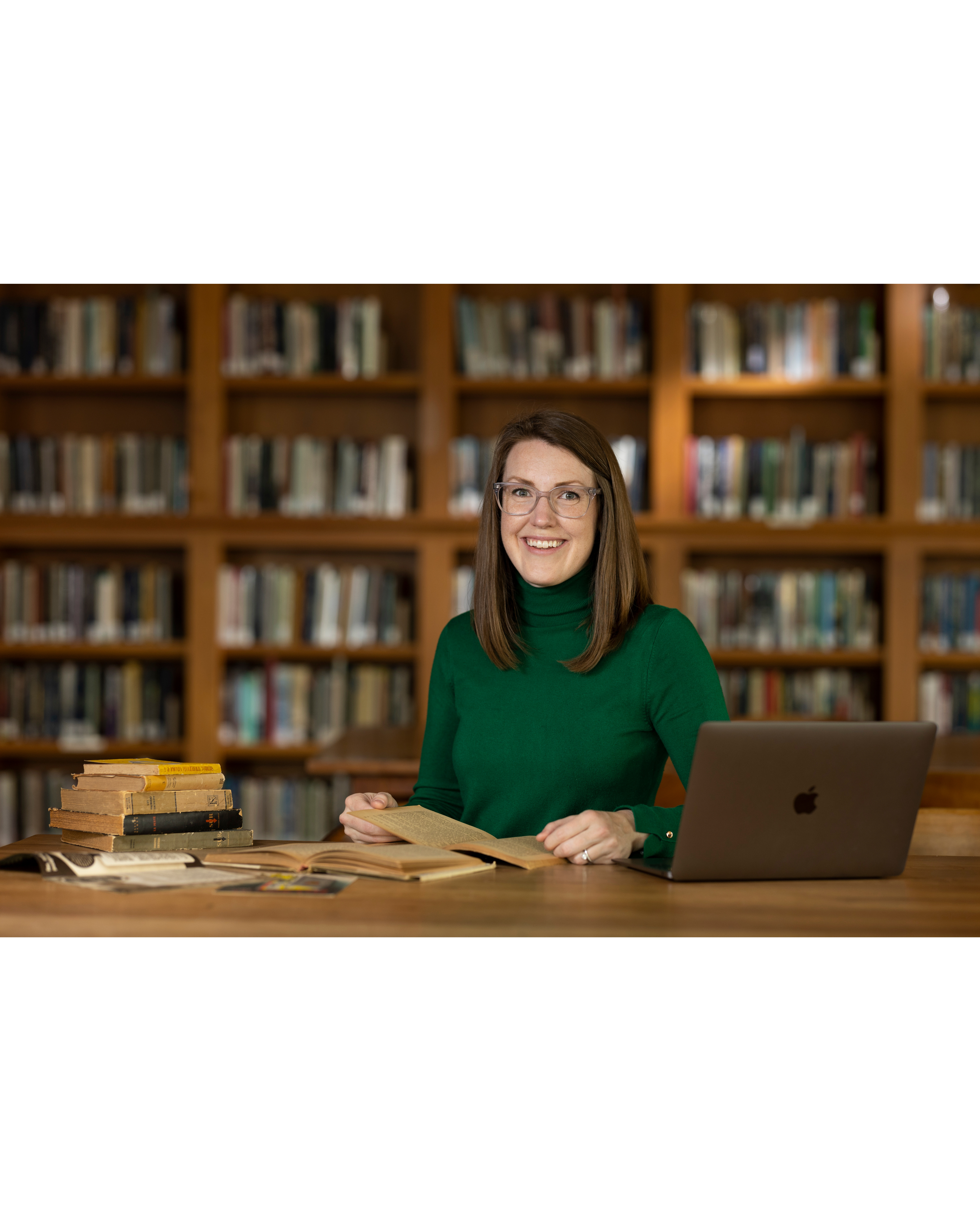 Photo of Nora Benedict, smiling with medium length brown hair and wearing a dark green turtle neck sweater and glasses, sitting at a desk with books and a laptop in front of bookshelves.