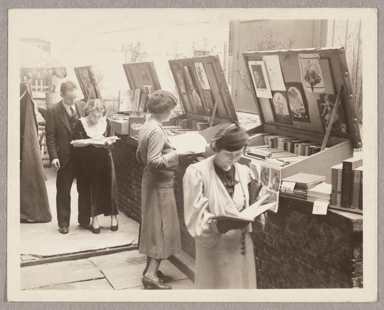 People browsing books at an outdoor bookstall with open boxes displaying books and prints.