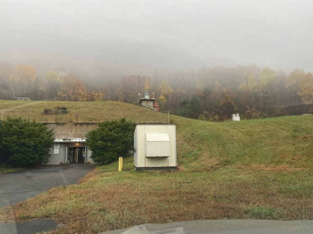 An outdoor scene of a partly underground facility. A grassy hill, possibly a bunker, blends into a foggy landscape with autumn-colored trees. A concrete entrance is visible at the base of the hill, flanked by two green bushes. In front of the entrance is a small, white utility shed with a sloped roof.