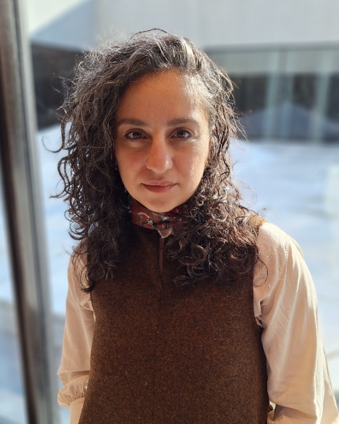 Photo of Michelle Al-Ferzly,  wearing a brown vest and a scarf and standing indoors near large windows.