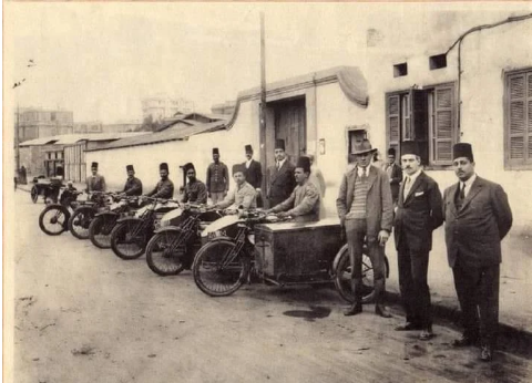 Sepia historical photograph of men posing with a row of early motorcycles outside a stucco building.